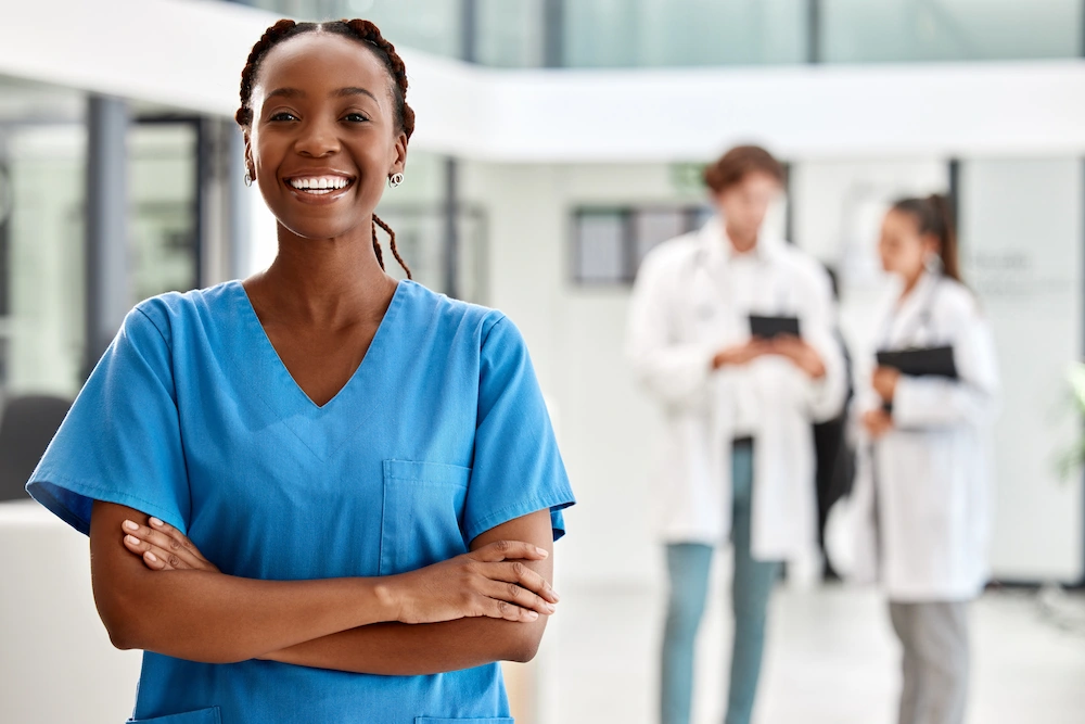 Smiling travel nurse standing with her arms crossed in a healthcare facility hallway.